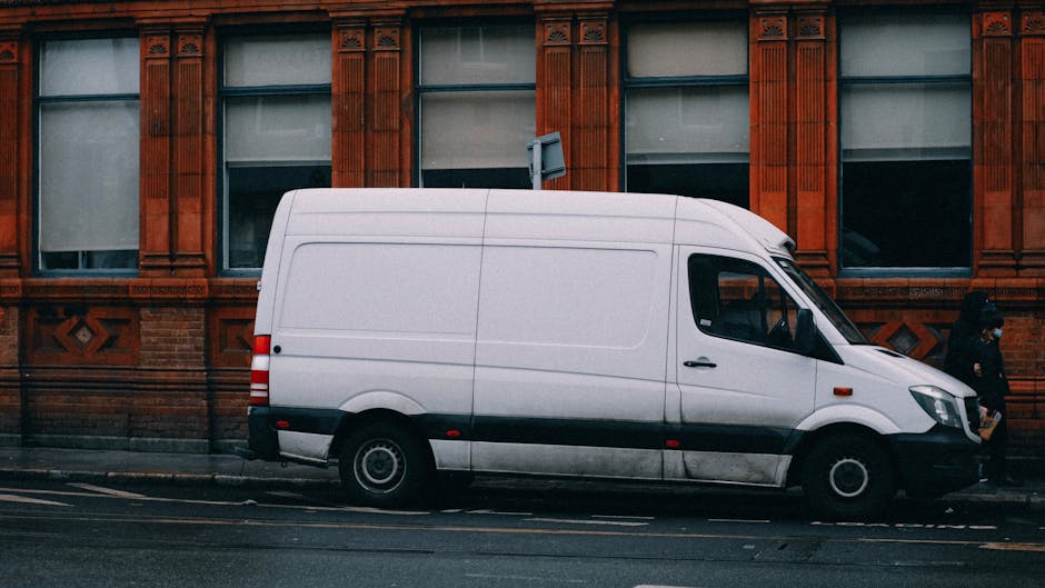 A man wearing a beige knit beanie, a green padded vest, a navy and grey athletic jacket, and black gloves is seated inside the open rear of a dark grey van, which is loaded with several medium-sized c
