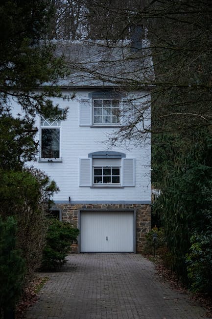 A three-storey residential house with a stone and white-painted exterior, featuring a white garage door at ground level and three windows with open shutters on the upper floors. The house is situated