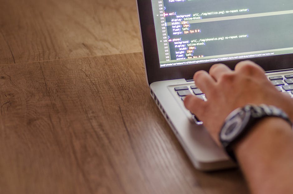 A person's hand, wearing a wristwatch, typing on a laptop keyboard placed on a wooden desk. The laptop screen displays lines of code with syntax highlighting, indicating activities related to digital
